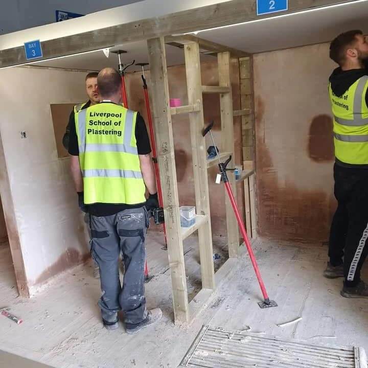 Students practicing plastering techniques at Liverpool School of Plastering, wearing safety vests in a workshop setting.